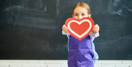 Portrait of adorable little girl holding red paper heart posing against blank blackboard in school