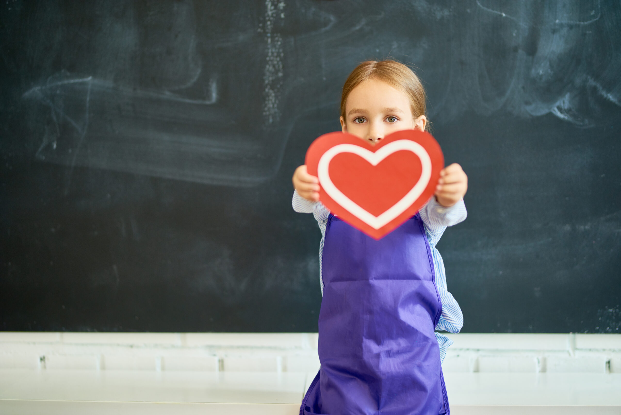 Portrait of adorable little girl holding red paper heart posing against blank blackboard in school