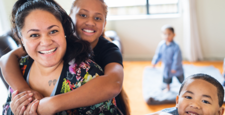 mother and her two children smiling at the camera, another child plays in the background
