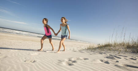 two young girls hold hands while walking on the beach