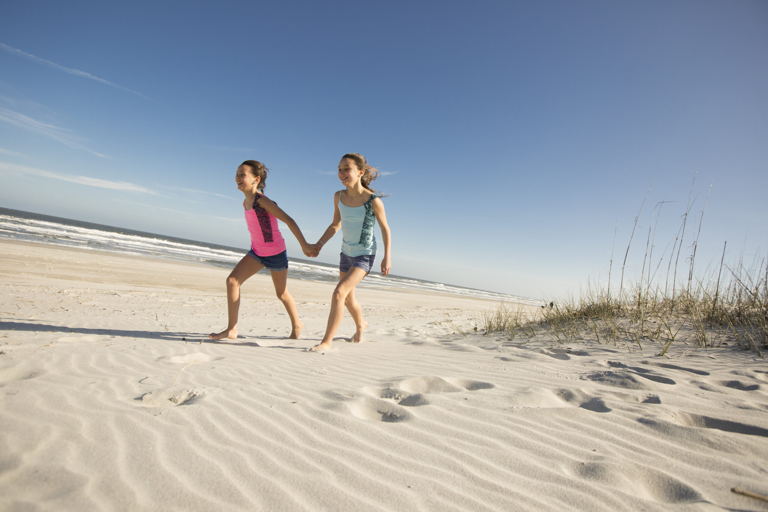 two young girls hold hands while walking on the beach