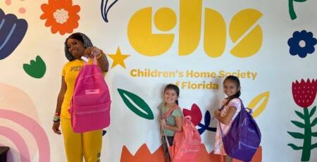woman and two young girls posing with new backpacks in front of a colorful wall that says CHS