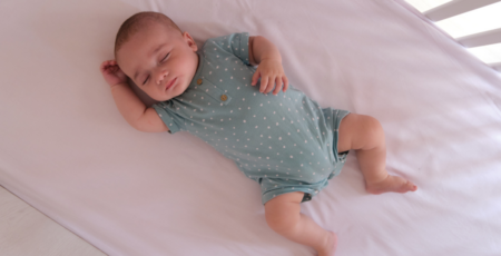 A baby sleeping on its back in a crib, wearing a light teal onesie with small white polka dots. The crib has a firm, flat mattress with a fitted sheet, following safe sleep guidelines.